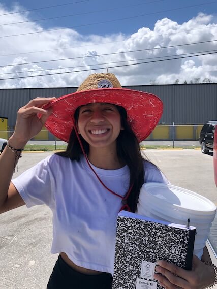 A photo of Elizabeth with a white shirt, wearing a straw hat, holding a black/ white composition book and with a big smile