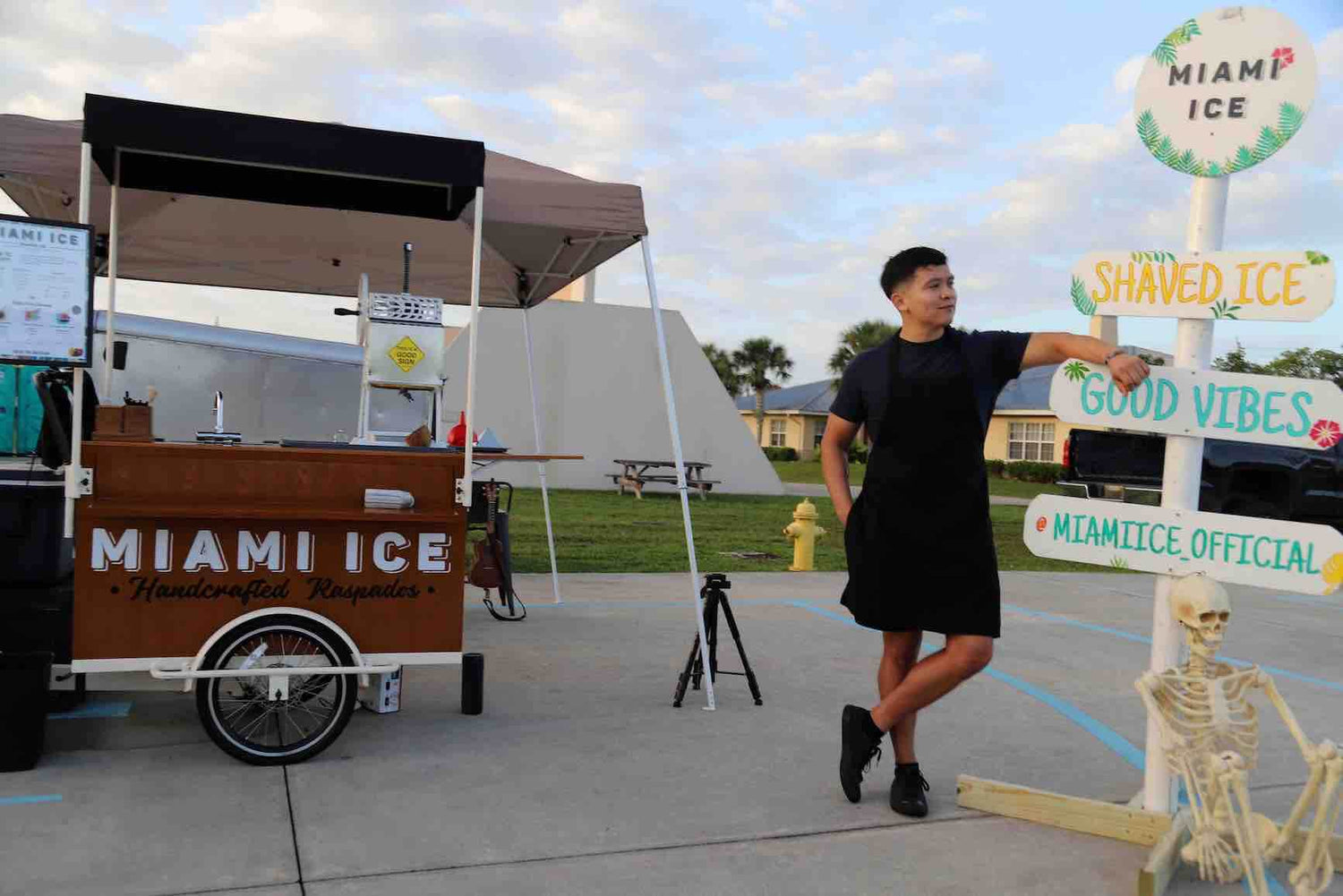 A picture of Joseph wearing a black apron and leaning against a white post that says "good vibes." Our Miami Ice bike cart is in the background on concrete and grass in the distance