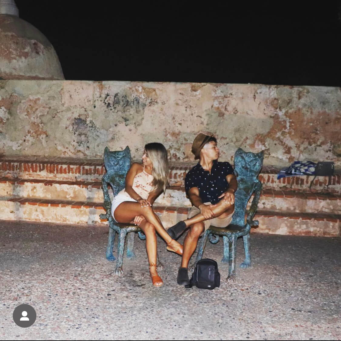 Two people (us) facing away from each other while sitting in metal chairs in the shape of cats. Old brick steps from a fort in puerto rico are behind us.